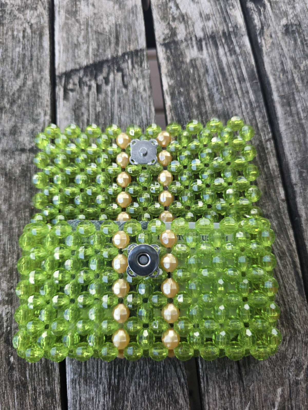 A handmade beaded wallet made of green faceted beads with vertical rows of opaque gold beads and two silver-tone magnetic button closures, displayed on a weathered gray wooden plank surface.
