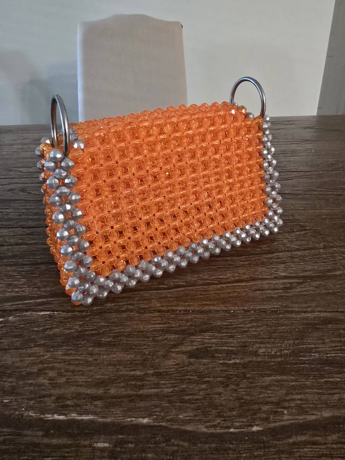 An orange beaded clutch bag with silver metallic bead borders and silver metal O-ring handles, resting on a dark wooden table.