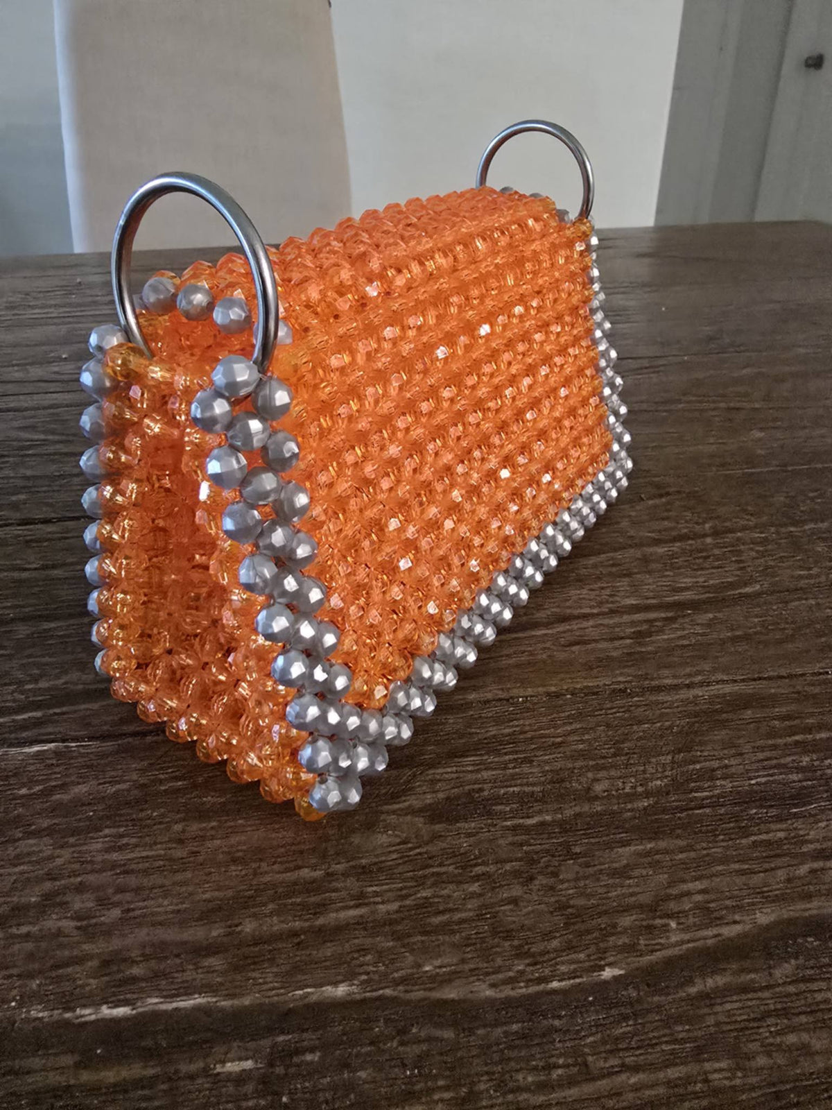 An orange beaded handbag with silver-tone metal D-rings and silver bead trim, resting on a dark wooden tabletop.