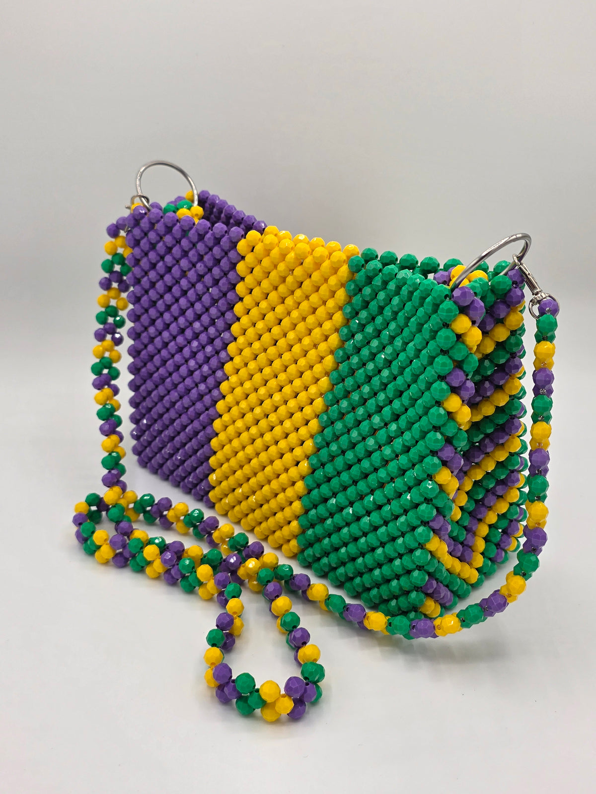 A beaded crossbody bag with a purple, yellow, and green vertical striped pattern, featuring a matching beaded strap and silver-tone metal rings. Perfect for Mardi Gras