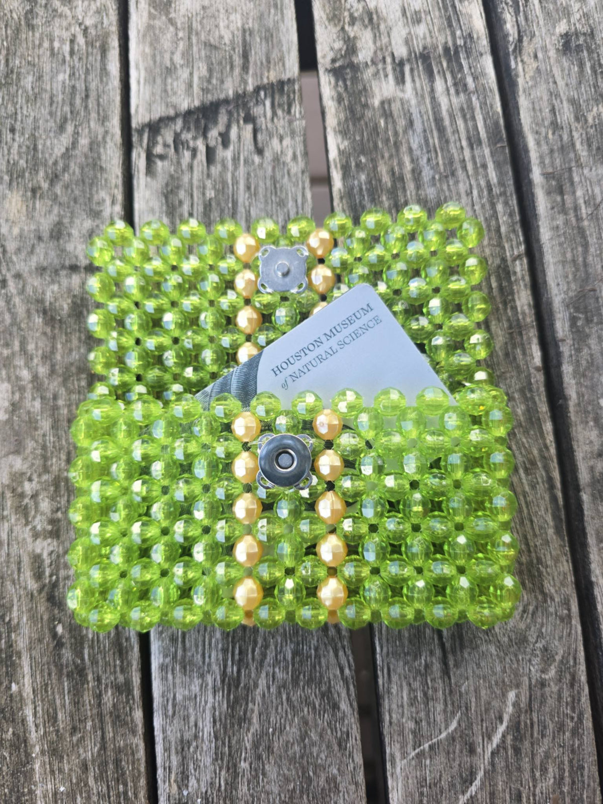 A handmade green beaded wallet with gold stripes and magnetic snap closures, shown on a weathered wooden plank surface with a paper tag attached.