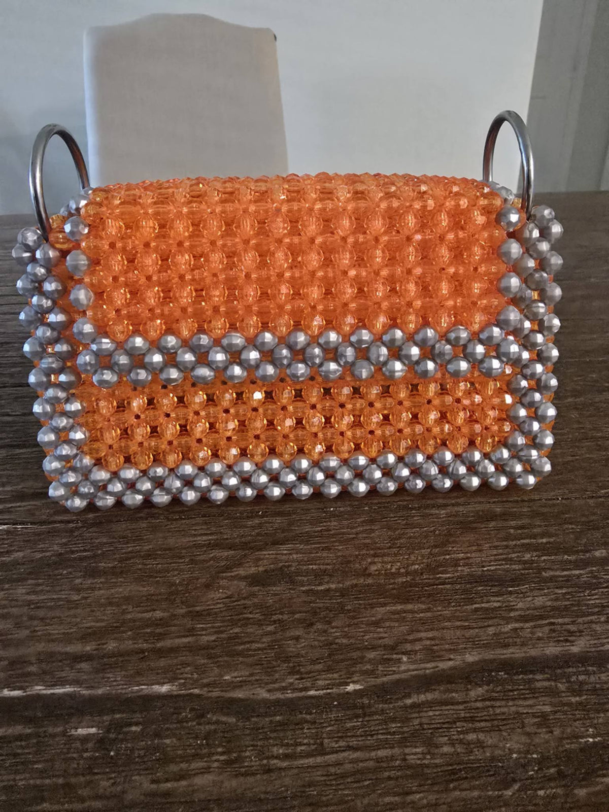 A handmade rectangular clutch bag made of orange and silver 8mm beads, featuring two silver metal O-ring handles, resting on a dark wooden table.