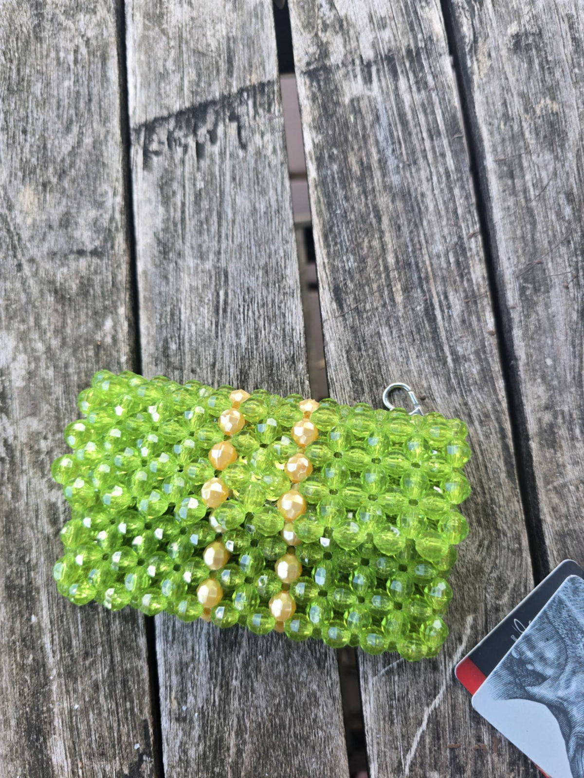 A handmade green beaded wallet with gold pearl-like beads and a silver keyring, resting on a weathered gray wooden plank surface.