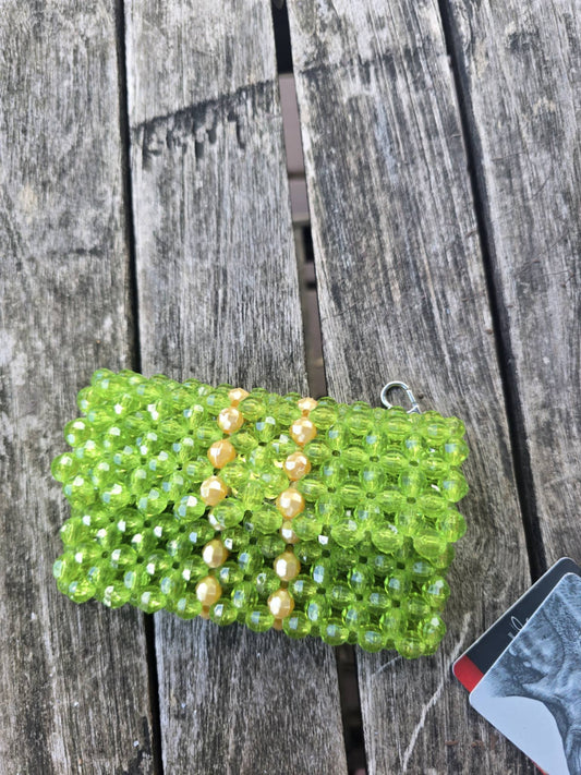 A handmade green beaded wallet with gold pearl-like beads and a silver keyring, resting on a weathered gray wooden plank surface.