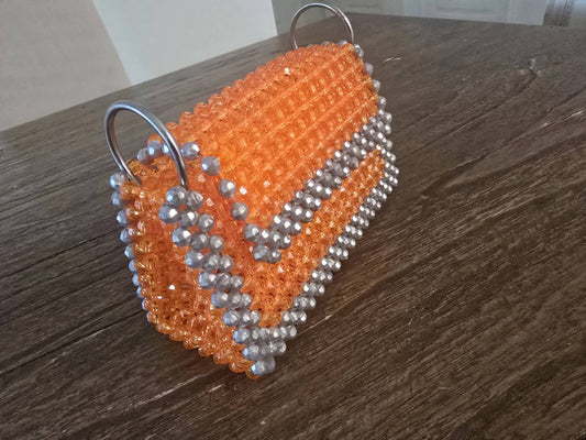 A handmade beaded handbag with a bright orange body, silver beaded trim, and silver metal D-rings, resting on a dark wooden table.