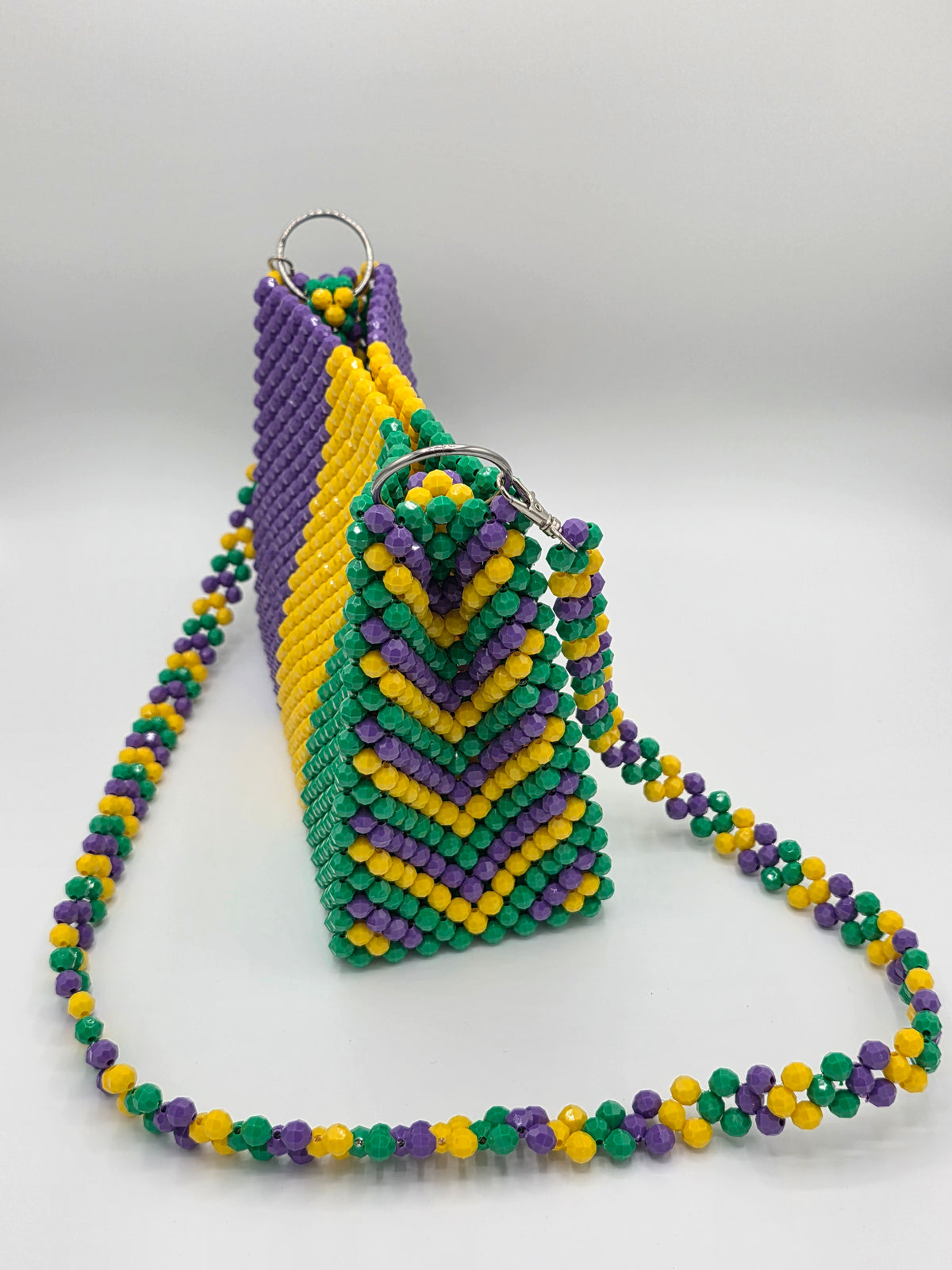 A beaded crossbody bag with a matching beaded strap, featuring a Mardi Gras color scheme of purple, green, and yellow. The bag has a geometric chevron pattern on the sides and is displayed on a light gray background.