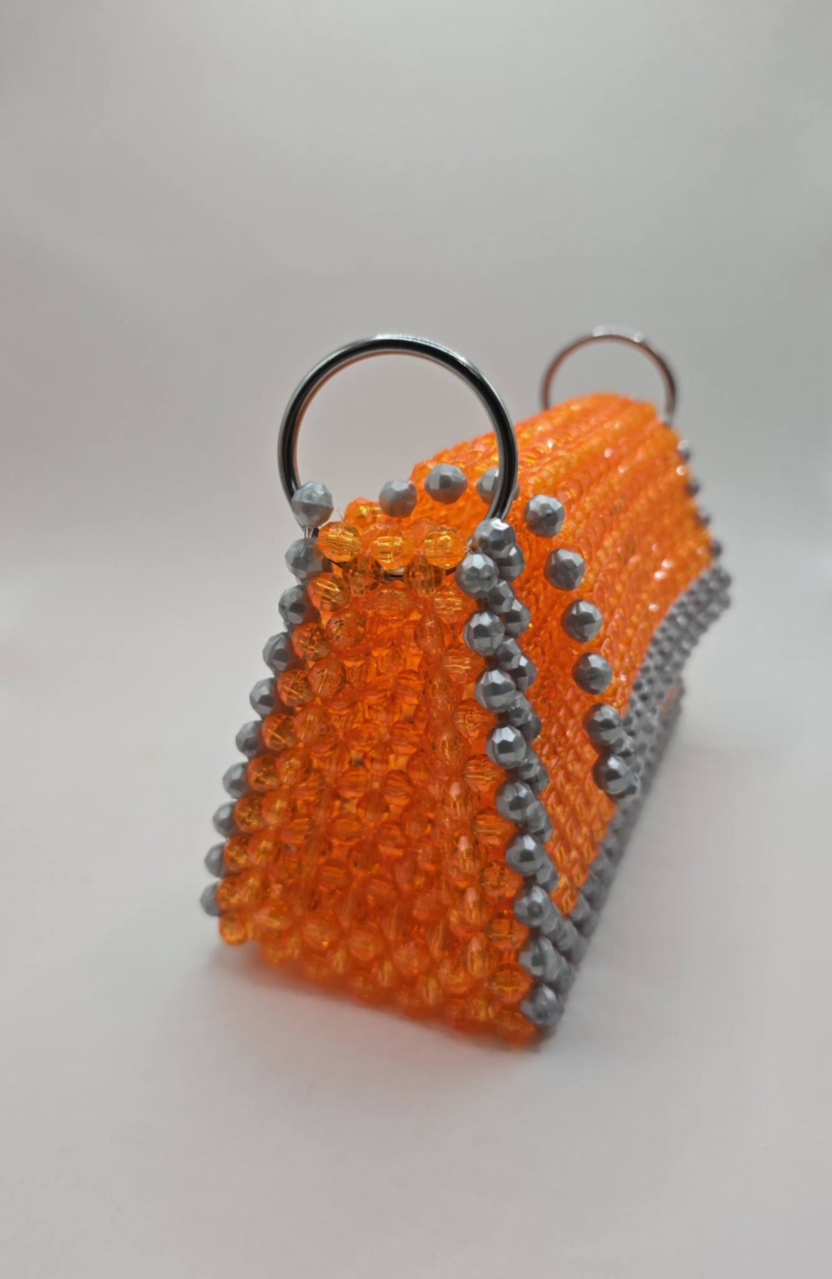 A small structured handbag made of bright orange faceted beads with silver-gray bead trim and two silver metal ring handles, photographed on a plain light background.