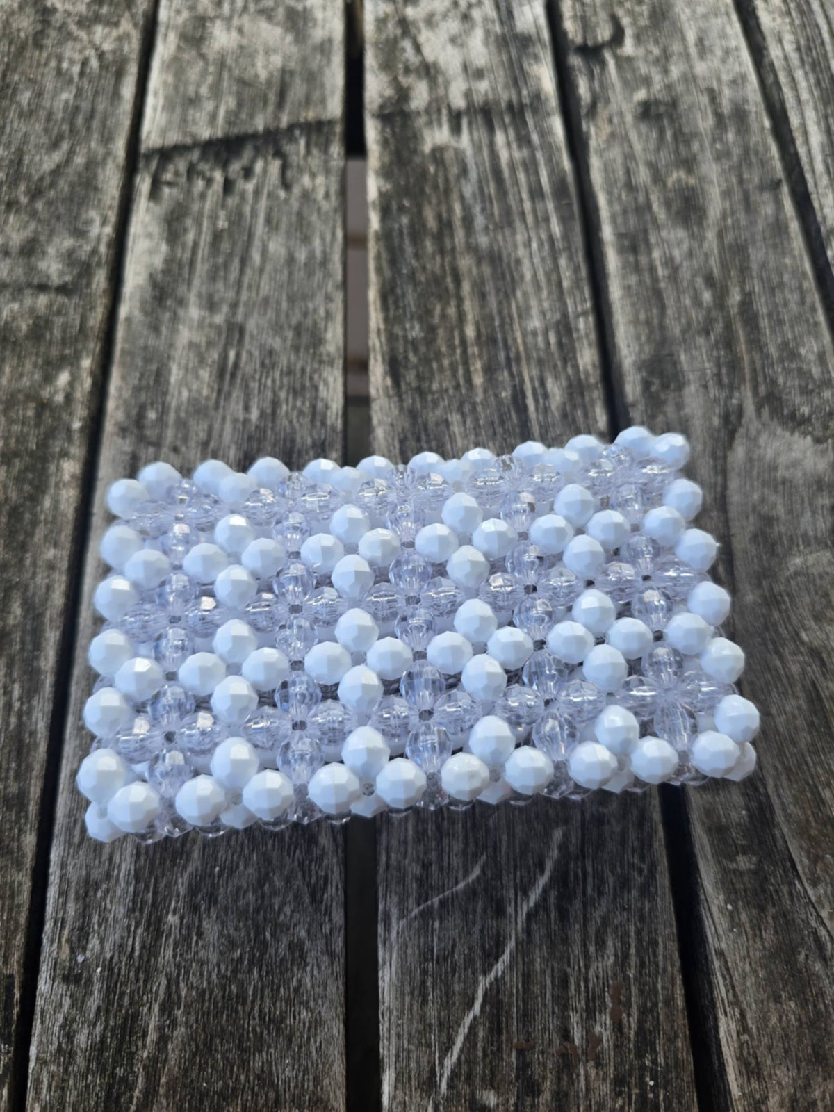 A handmade wallet, covered in white faceted beads with a clear beaded border, placed on a weathered gray wooden plank surface.